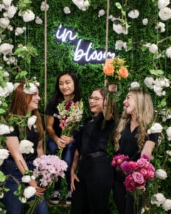 4 In bloom employees smiling and posing with flowers in front a a green backdrop with white flowers with a neon sign that says In Bloom
