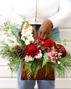 A hand holding a Petals and Presents Arrangement. The flower arrangement is CHristmas greenery, red and white flowers in a basket the shape of a purse